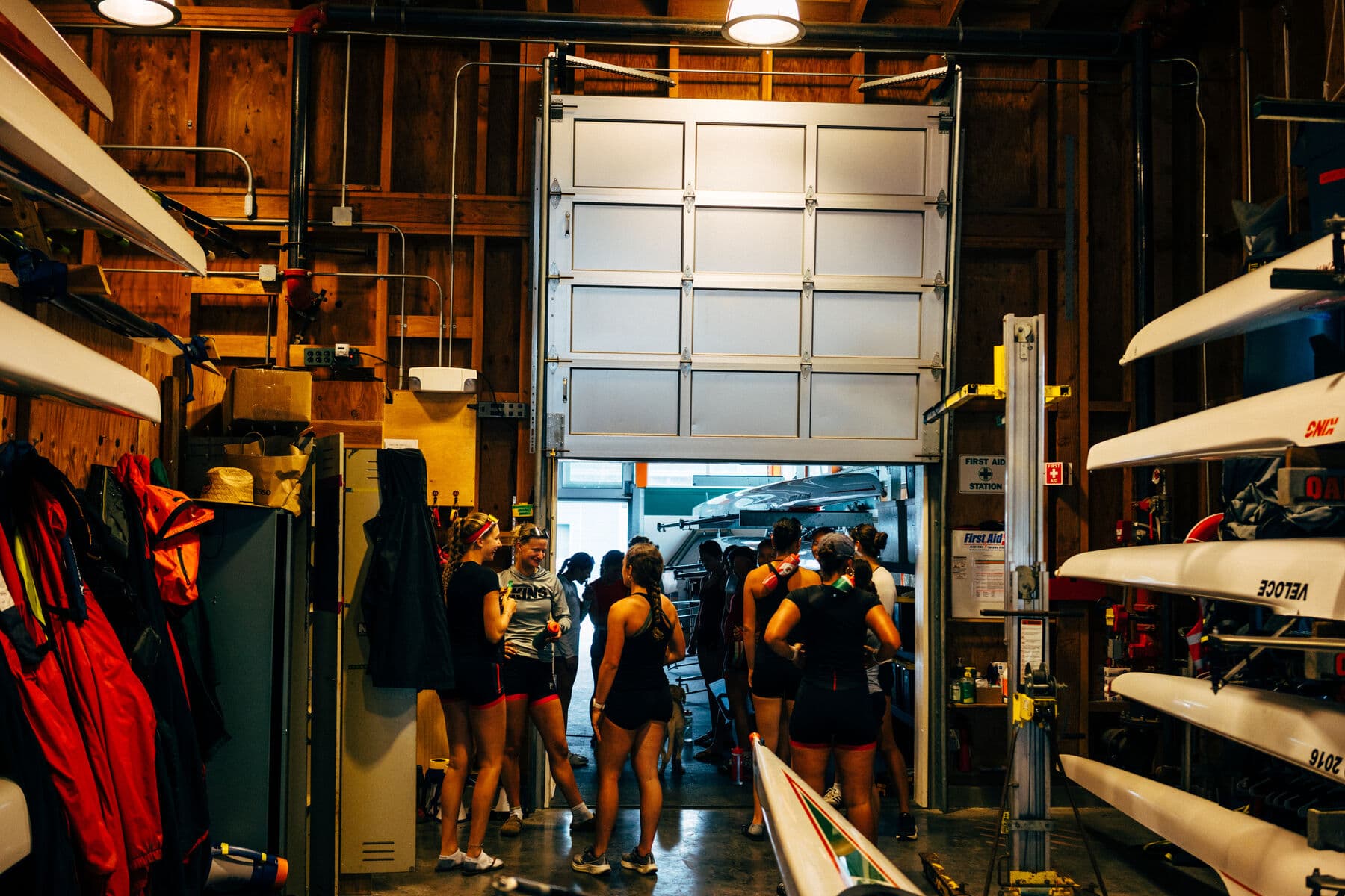 Athletes gathered in the doorway of a boat bay, racked eights and quads on both sides