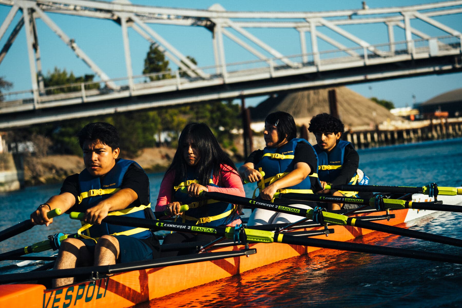 Four young rowers pull together in an orange training boat under a bridge on the Oakland Estuary