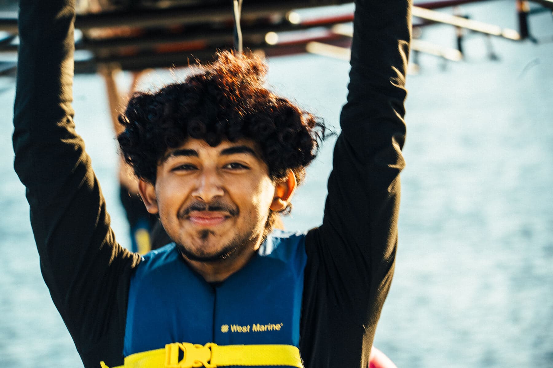 A young rower in a life jacket smiles with his arms raised overhead on the dock