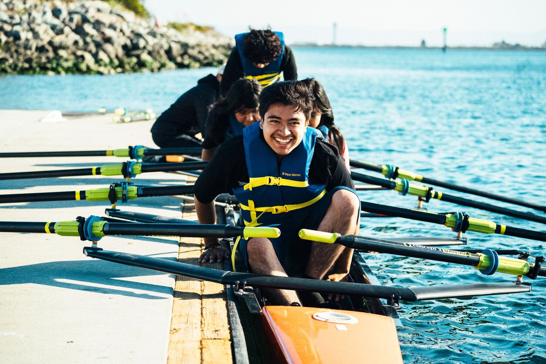 A young rower grins from his seat as his crew settles into the boat at the dock