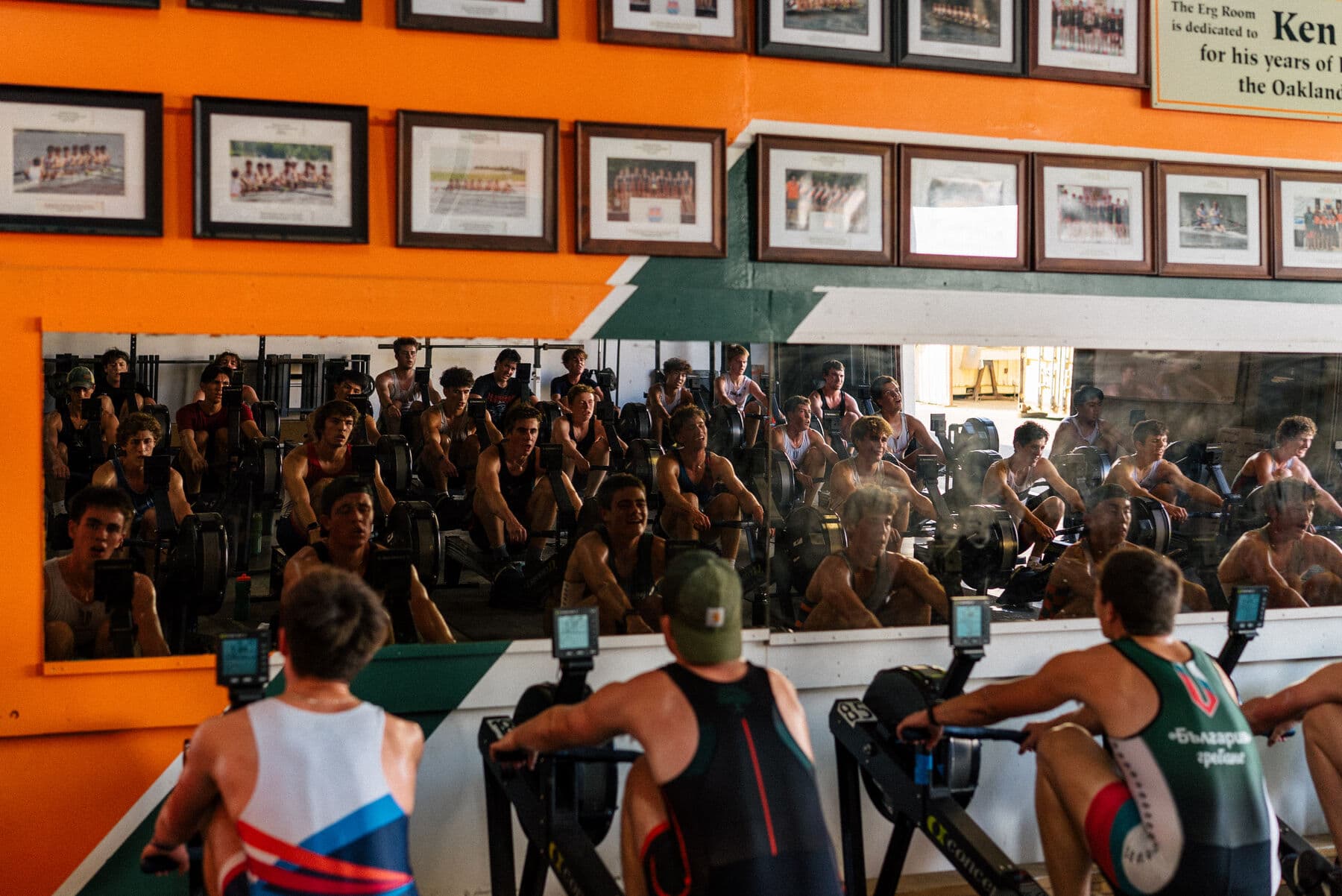The high school team fills the same erg room, rows of ergs reflected in the mirror under the photo wall
