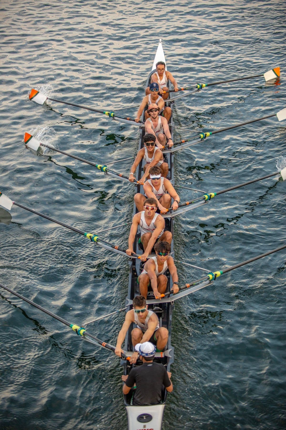A men's eight from above, rowers in white OAKLAND tanks, the coxswain at the stern
