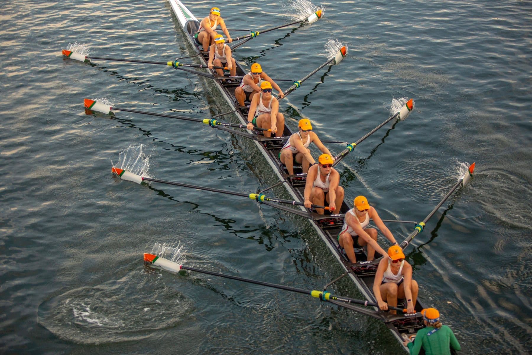 The women's eight in orange caps seen from directly above, blades squared and spray flying