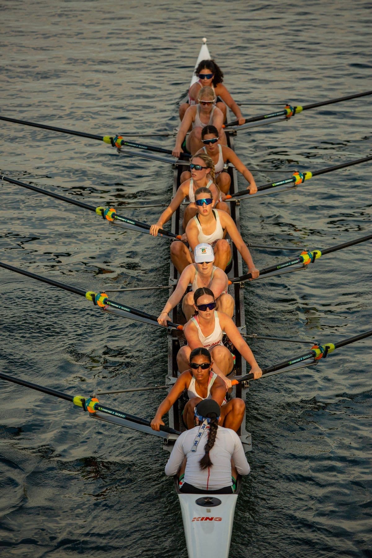 A women's eight from above, every rower reaching forward together, the coxswain at the stern