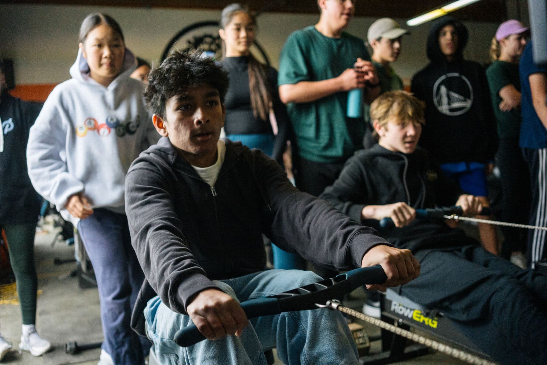 A middle schooler pulls through an erg piece in the boathouse while teammates watch