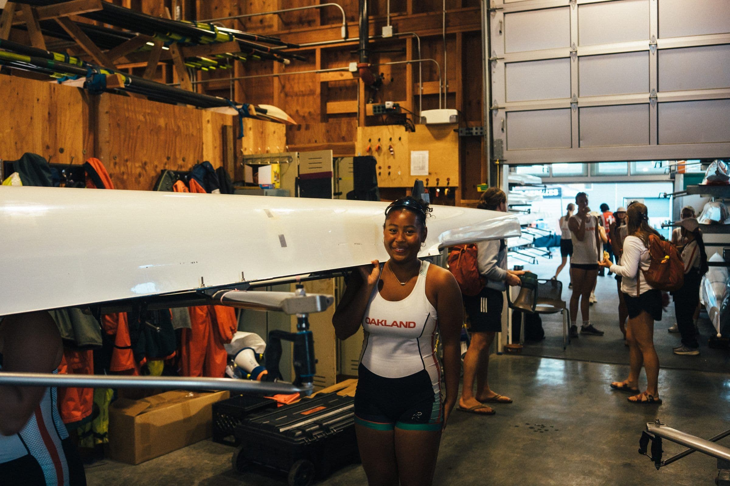 A rower smiles as her crew carries their boat out of the boathouse