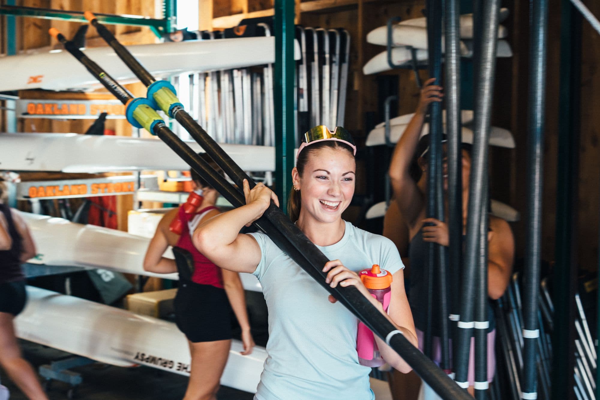 A rower laughs as she carries oars out of the boathouse before practice