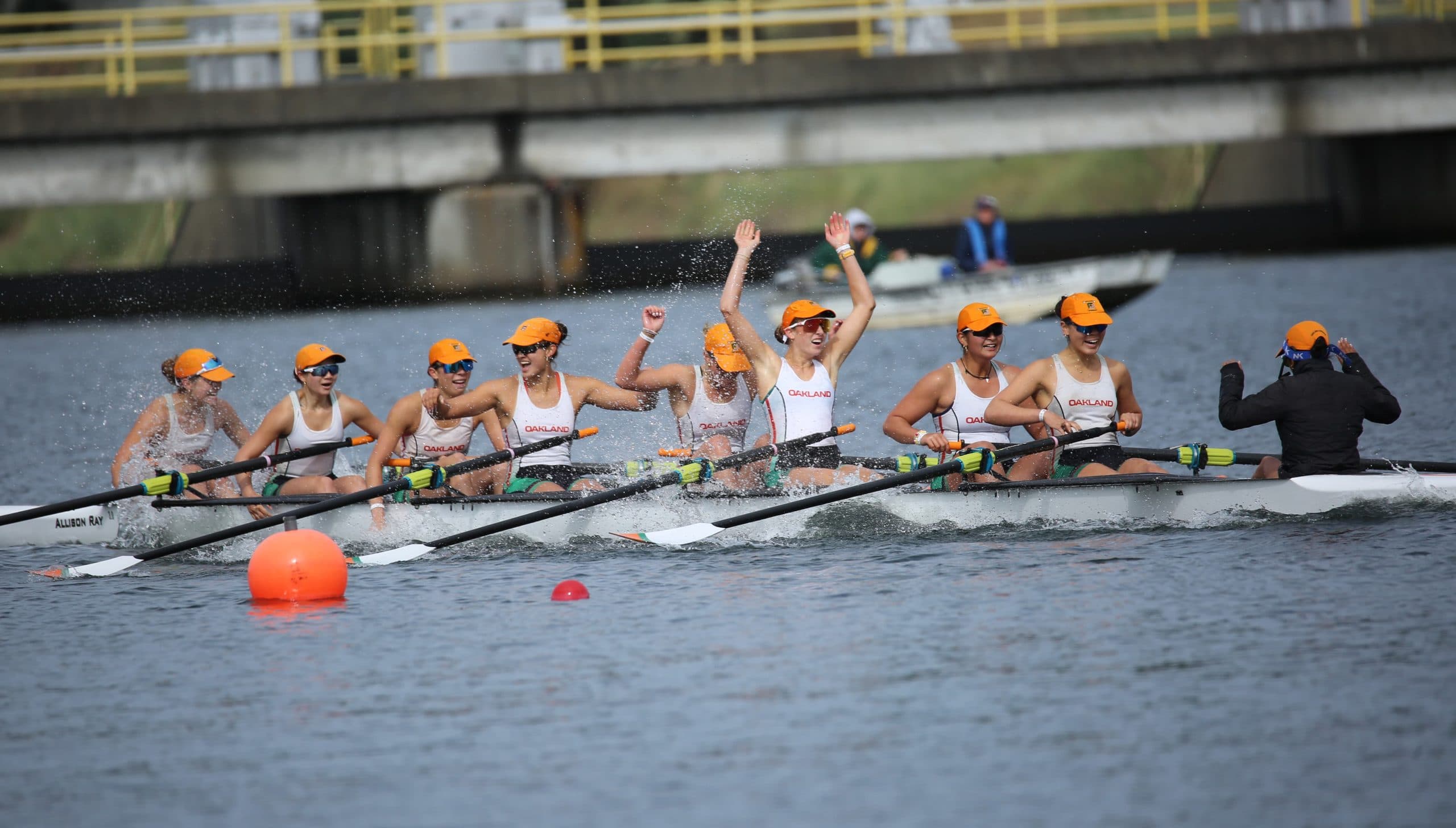 The women's eight crosses the line with arms raised, orange caps and spray flying