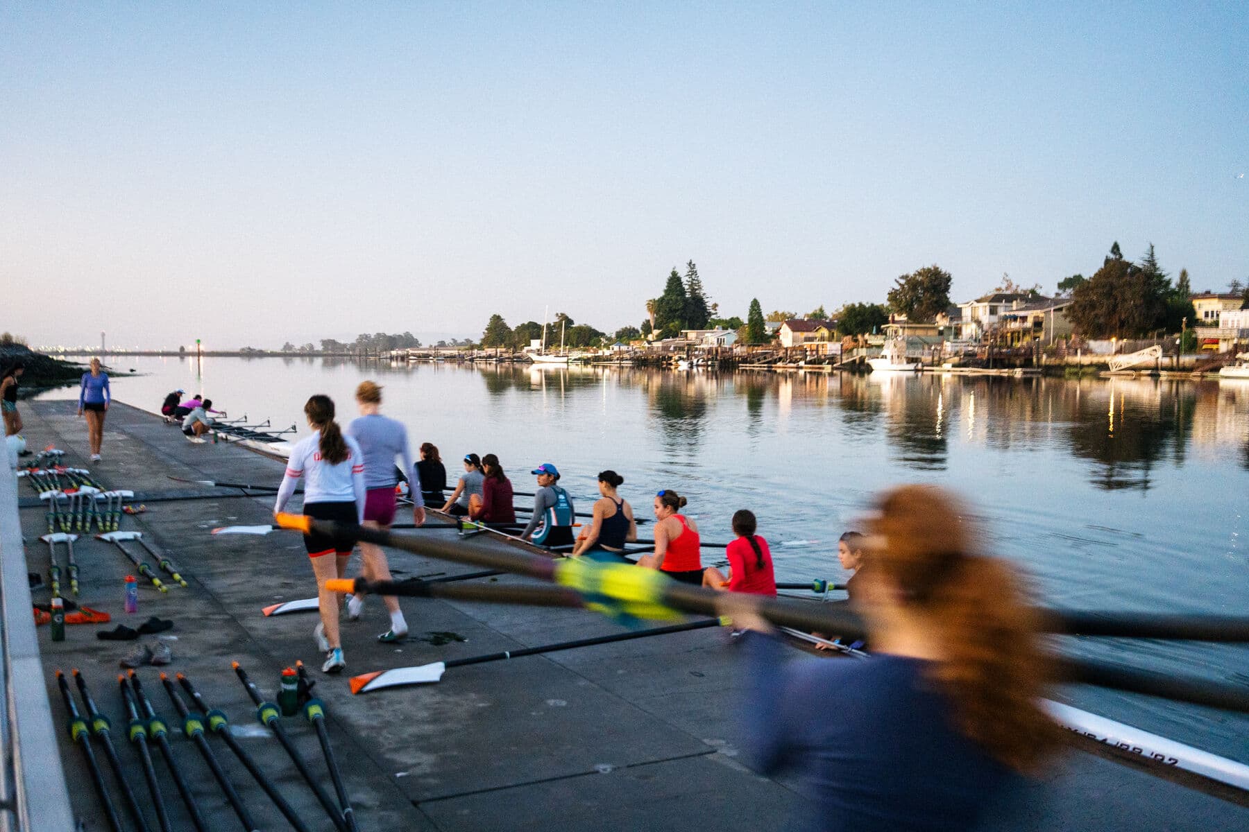 The women’s squad carrying a boat down the Tidewater dock, oars laid out ready, the Estuary houses across the water
